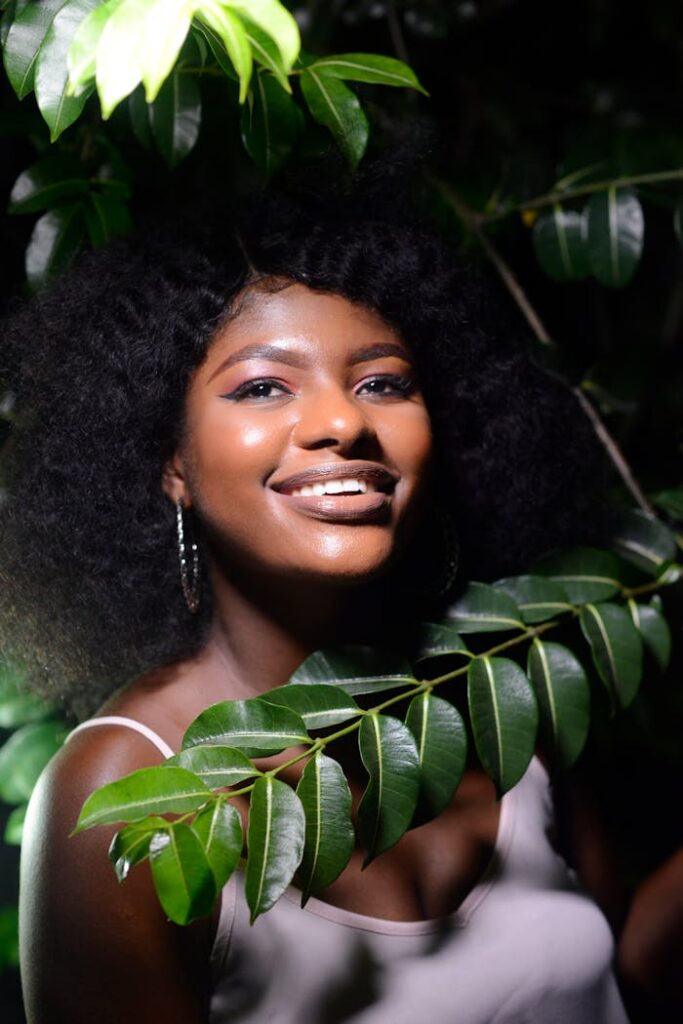 Smiling woman with curly hair and earrings posing amidst lush green leaves outdoors.