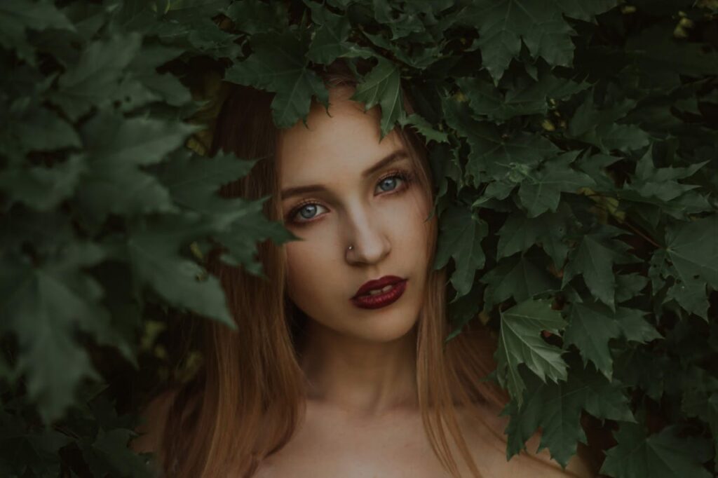 Portrait of gentle female model with red lips looking at camera while touching maple leaves with head