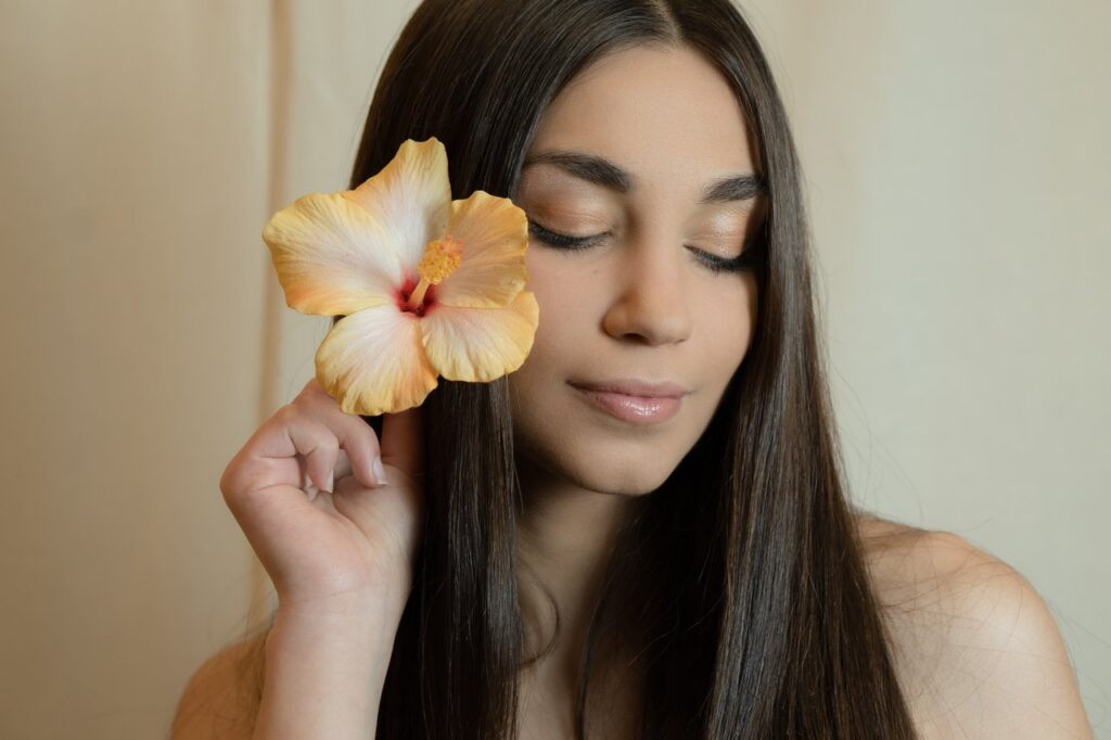 Close-up portrait of a serene woman holding a hibiscus flower against her face, eyes closed, indoors.